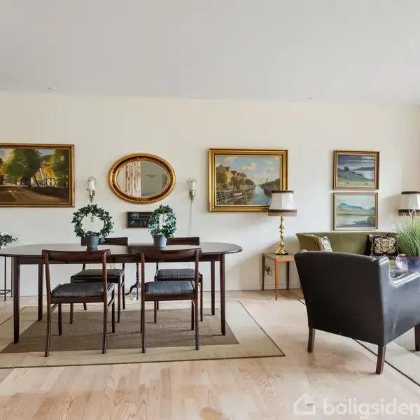 Spisestuemøbler står på et tæppe i en stue med parketgulv. Tre malerier og et spejl hænger på væggen. Der ses en lænestol og sofabord ved siden af.