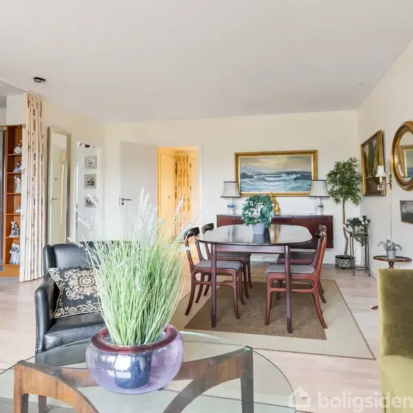 En stue indeholder et sofabord med en plante i forgrunden, omgivet af stole og en sofa. I baggrunden er der en spisestue med et bord og stole, kunst på væggene og en åbning til et køkken.