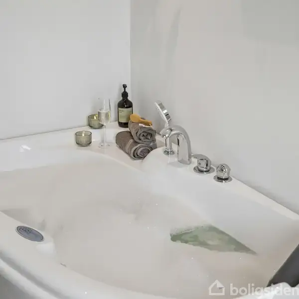 Badekar med skumfyldt vand, omkranset af sammenrullede håndklæder, stearinlys og flasker. Badekarret er placeret i et lyst badeværelse med hvide vægge.