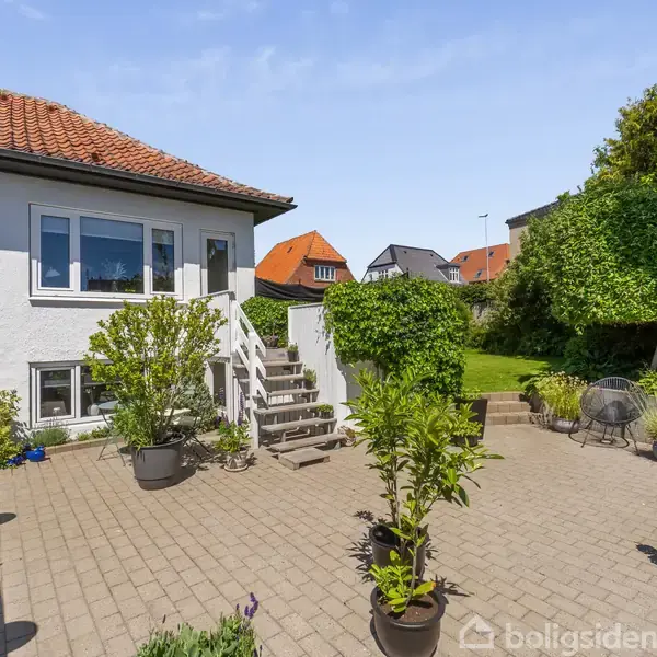 Et hus med hvid facade og rødt tegltag. En stenterrasse med blomsterkrukker omkranser området. Trappe fører op til indgangen. Omgivet af andre huse og grøn beplantning.