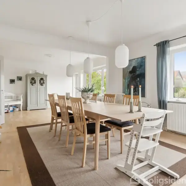 Spisebord med træstole står i en lys spisestue med store vinduer, der lukker dagslys ind. Et skab, lysarmaturer og grønne planter dekorerer rummet.