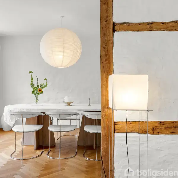 Et spisebord med hvide stole er omgivet af en rustik hvid væg med trækonstruktioner. En kuglerund loftslampe hænger over bordet, som har en vase med blomster.