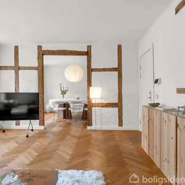 TV står på stativ ved siden af stort vindue. Spisestue kan ses gennem åbning med synlige bjælker. Trægulv og moderne skænk skaber en elegant atmosfære.