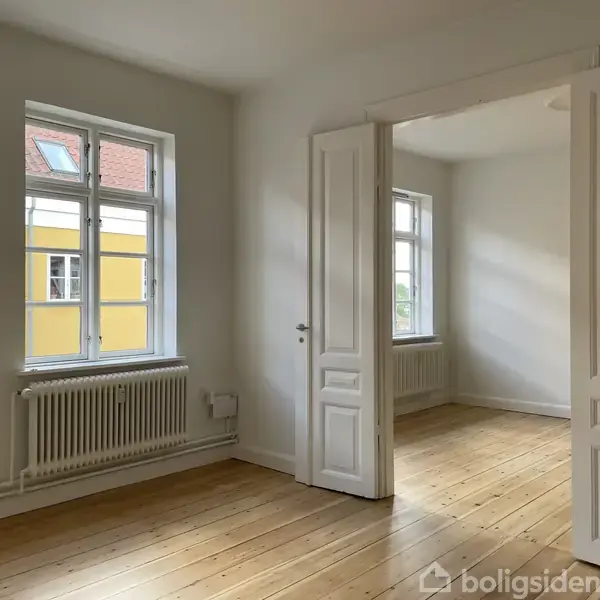 Et lyst, tomt værelse med lyse trægulve og store vinduer. Døre åbner til et tilstødende rum, og der er en radiator under vinduet. Udenfor ses en gul bygning.