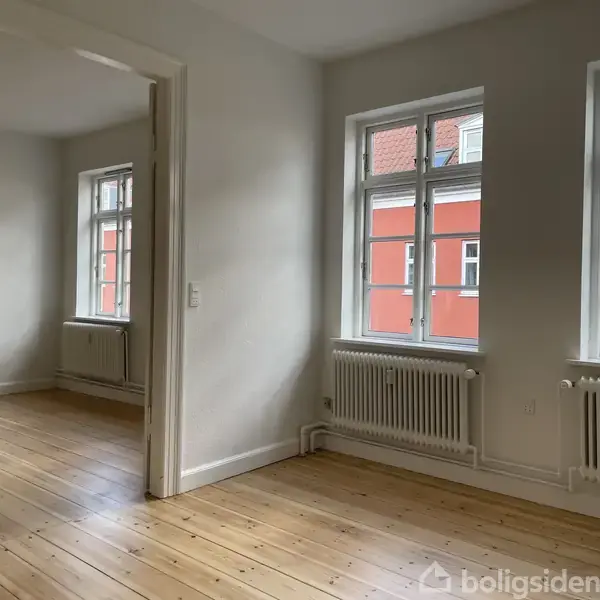Et tomt værelse med trægulv og hvide vægge har to store vinduer med udsigt til farverige bygninger. Radiatorer er placeret under vinduerne.