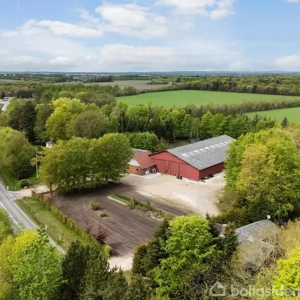 Rødt lagerbygning står stille omgivet af grønne træer. Bygningen ligger ved en vej, der snor sig gennem et frodigt, landligt landskab med marker i baggrunden.