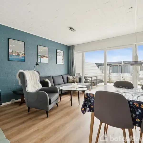 Stue med sofagruppe og spisebord. Sofabord og stol står foran en blå væg med væghængte billeder. Store vinduer med udsigt til terrasse og blå himmel.