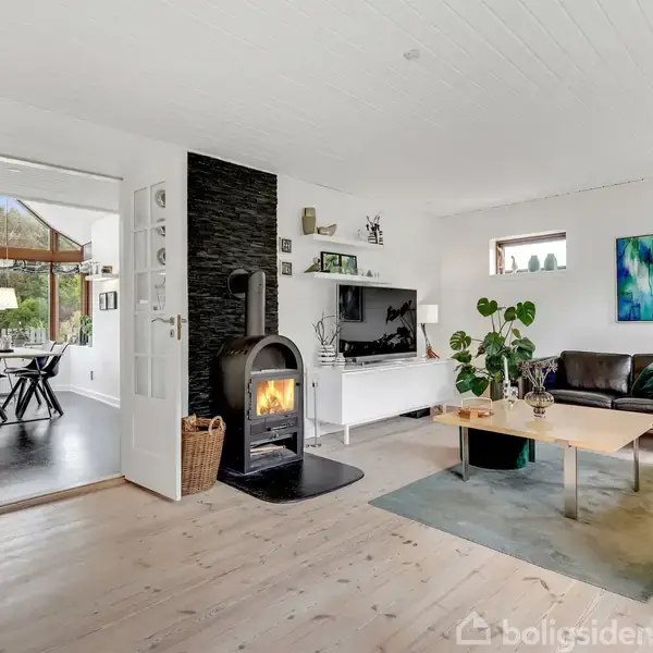 Stue med brændeovn ved væg, sofa og sofabord, stor plante. Spisestue i baggrunden med spisebord og stole. Hvide vægge, træloft og parketgulv, kunst på væggen.
