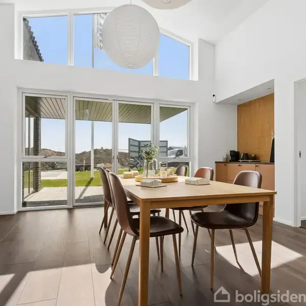 Spisebord med seks stole står på et trægulv i en lys spisestue. Store vinduer med udsigt til en terrasse og grønne områder. Minimalistisk og moderne indretning.