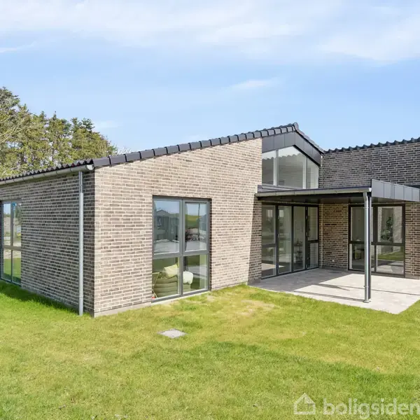 Et moderne etplanshus i grå mursten med store vinduer og fladt tag står i en grøn have omgivet af græsplæne. Skov i baggrunden under en lys, blå himmel.