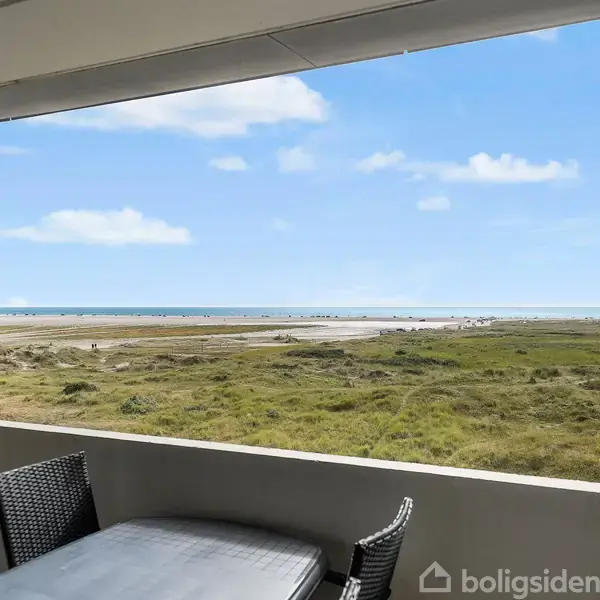 Bord omgivet af kurvestole står på en altan. Udsigt over en grøn slette strækker sig ud mod en strand med havet i horisonten under en blå himmel.