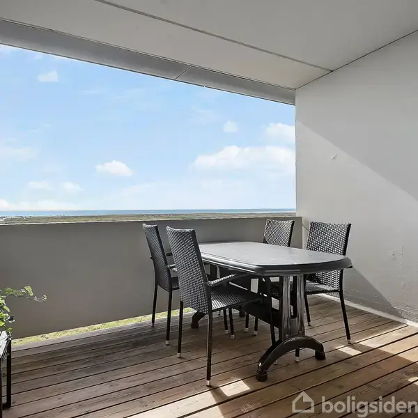 Terrasse med havemøbler; et bord og fire stole står på trædæk. En plante i potte til venstre. Rummet er delvist overdækket med udsigt til blå himmel og skyer. Ingen tekst.