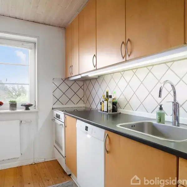 Et køkken med træskabe og sort bordplade; indeholder en vask og opvaskemaskine. Flisevæg og stort vindue med planter i baggrunden giver lys og rummelighed.