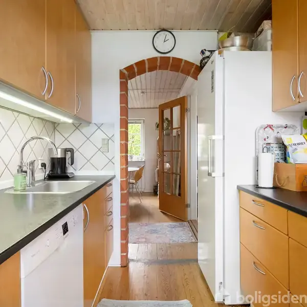 Et smalt køkken med træskabe og sorte bordplader. En kaffemaskine står på bordet ved siden af en opvaskemaskine. En bueformet døråbning fører til en lys stue. En vægur hænger over døråbningen.