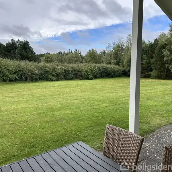 En terrasse med kurvemøbler står stille ved et bord på fliser. I baggrunden kan ses en stor, velplejet græsplæne omgivet af høje buske og træer under en blå himmel.