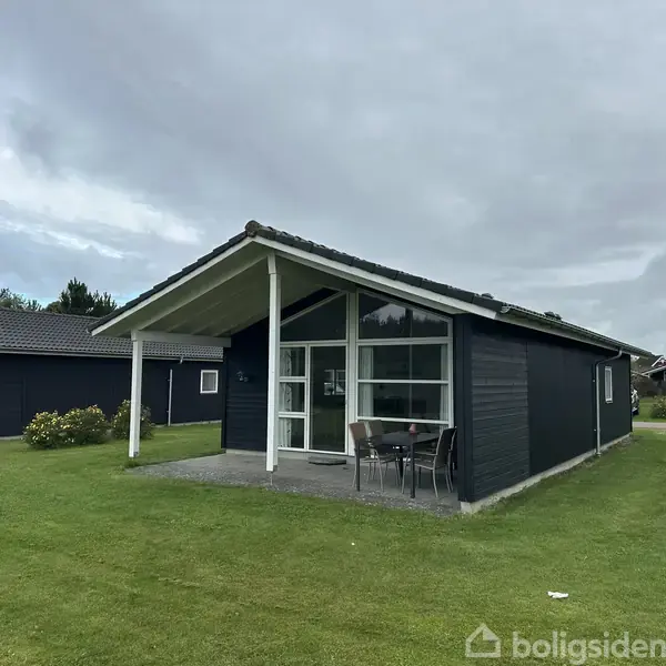 Et sort træhus med store vinduer står på en grøn græsplæne. Foran huset er et lille terrasseområde med et bord og to stole.