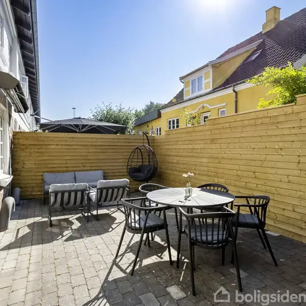 En udendørs terrasse indeholder et rundt bord med fire stole og en sofa, omgivet af et træhegn. I baggrunden ses en gul bygning og en klar blå himmel.