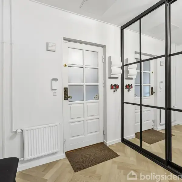 Dør står ved siden af en stor væg med spejle, mens rum med hvidmalede vægge og parketgulv rummer sort knagerække og radiator.