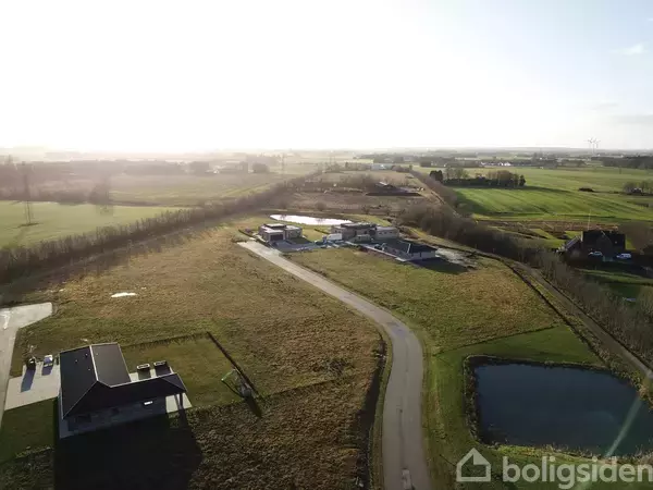 En landejendom med moderne bygninger omgivet af åbne marker og en snoet vej. En lille dam er placeret til højre i forgrunden under en solbelyst himmel.