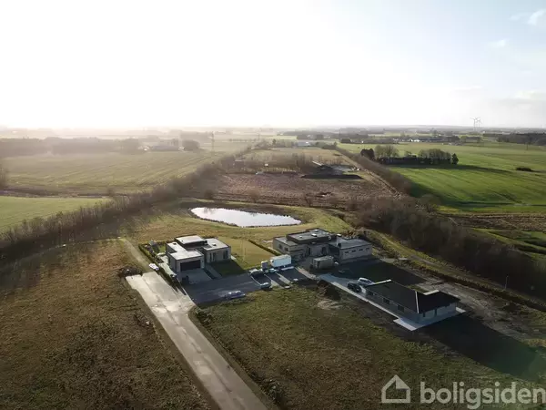 Huse står stille omgivet af marker og en mindre sø i en landlig, solbeskinnet landskab.