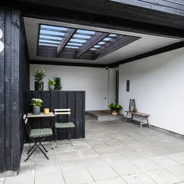 En sort og hvid, overdækket terrasse med nummeret "3" på væggen. Der er et lille bord med stole og planter i krukker samt en bænk i baggrunden.