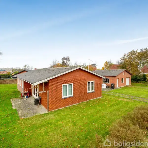 To røde murstenshuse står på en grøn græsplæne med træer og buske omkring. Himlen er klar blå, og landskabet er åbent med flere træer i baggrunden.