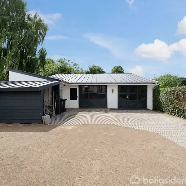 Et moderne, hvidt hus med fladt tag står stille i en have. Ved siden af er en sort garage. Omgivet af hække og grønne træer under en blå himmel.