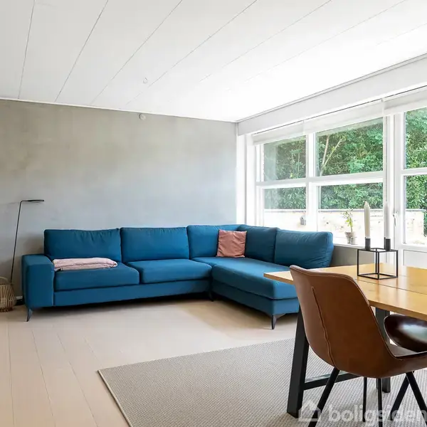 Blå hjørnesofa står foran en stor vinduesfront, omgivet af indendørs planter og et spisebord med brune stole. Rummet er lyst med en minimalistisk indretning og udsigt til have.