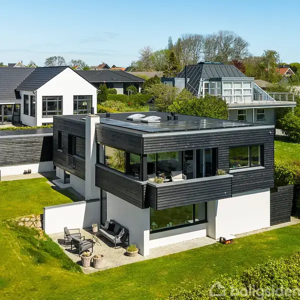 En moderne, to-etagers bolig står omgivet af velplejede grønne haver i et villakvarter. Huset har store vinduer, fladt tag og terrasse med havemøbler på bagsiden.