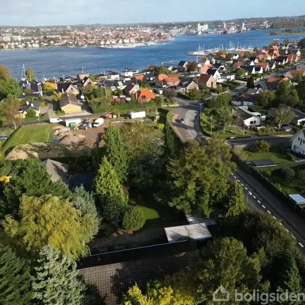 Et boligkvarter med huse og træer; det ligger tæt ved en bugt med en by i baggrunden, hvor mange bygninger er synlige i horisonten under en klar himmel.
