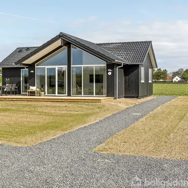 Et sort træhus med store vinduer står på en grussti. Der er en terrasse med havemøbler. Omkring huset er der en græsplæne i et landligt område.