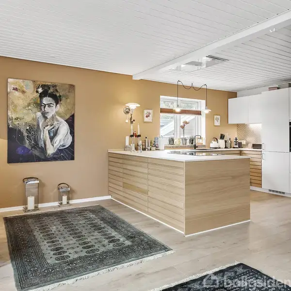 Et moderne køkken med træskabsø, set fra stue med tæpper og kunstværk på væggen. Rummets lyse farver og vindue giver naturligt lys. Bord med bøger og lampe ved siden af.