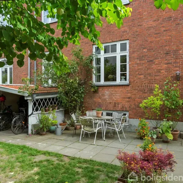 En gårdhave med havemøbler står på en flisebelagt terrasse ved en murstensbygning. Cykler hviler under et skur ved siden af. Der er planter i krukker omkring.