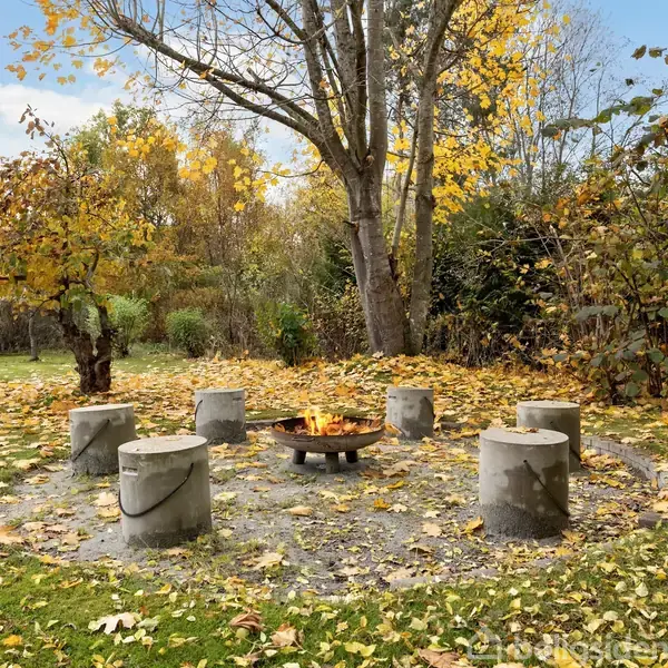Bålplads står i centrum, omgivet af fem stensæder. Træer med gule og grønne blade omringer området, som er dækket af visne blade. Det er udendørs i en have.