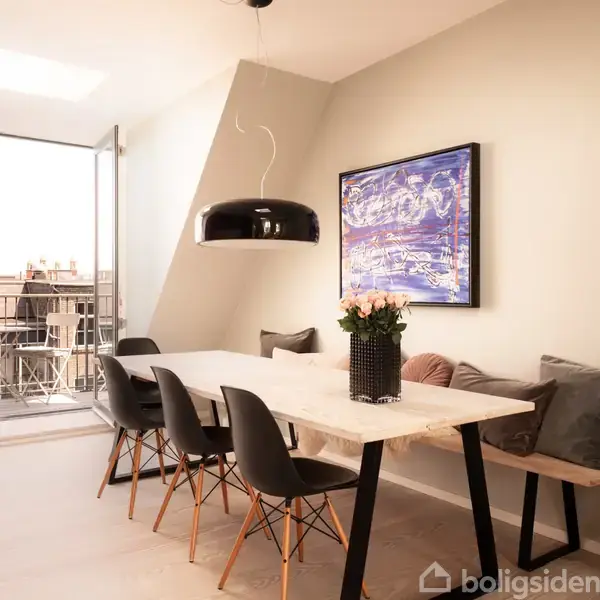 Et spisebord er omgivet af sorte stole i en lys stue med åbne altandøre. En sort lampe hænger over bordet, og der hænger et maleri på væggen. En balkon er synlig udenfor.
