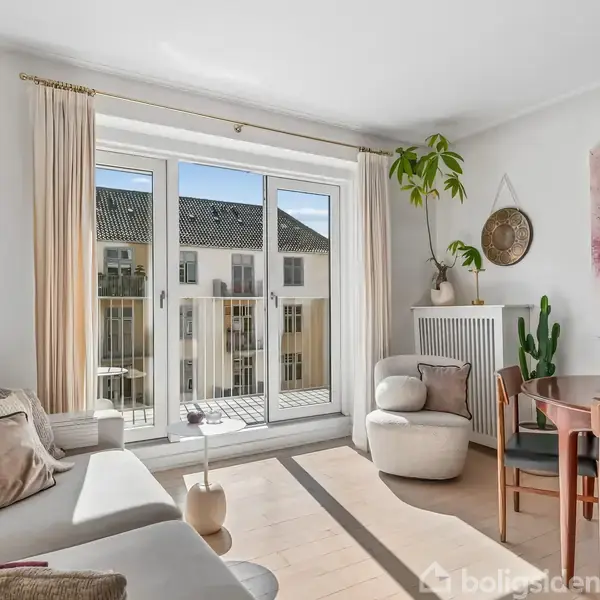 En stue indeholder en hvid sofa og spisebord, planter og lysindfald fra store vinduer. En altandør fører ud til en udsigt over bygninger. Møblerne er lyse og moderne.