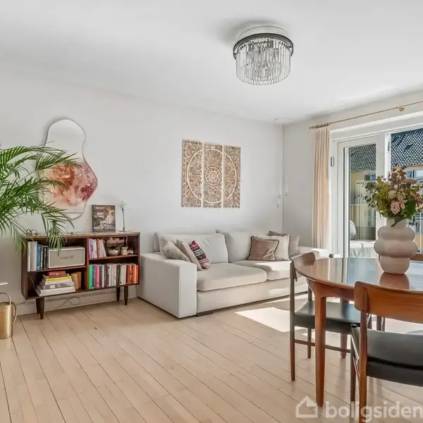 Sofa er centralt placeret i en lys stue ved siden af et stort vindue med skydedør. Rundt bord med stole, bogreol og plante. Vægdekorationer over sofaen.