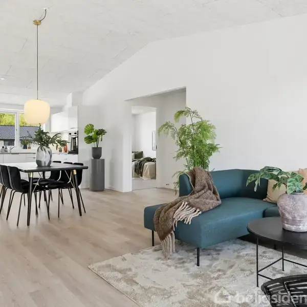 Stue med blå sofa, dekoreret med planter og tæppe, ved siden af rundt sofabord. Baggrunden viser køkkenalrum med spisebord og stole, hængende lampe, vinduer med udsigt til haven.