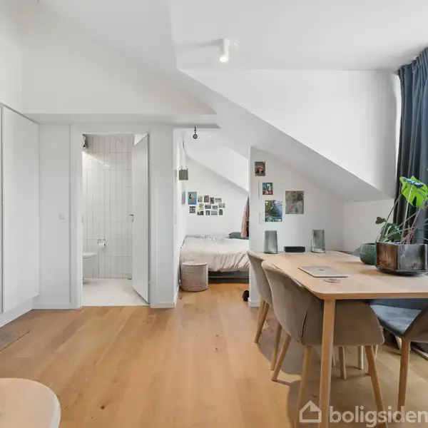 En lys stue med trægulv, spisestuebord og stole. Store vinduer med blå gardiner giver udsigt til bygninger. En plante står på bordet; bagved ses en seng og badeværelse.