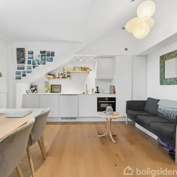 En lys stue/køkken kombination med trægulv; sofa til højre, spisebord til venstre. Køkken med åben hylde over. Billedvæg ved spisebordet, spejl over sofaen.