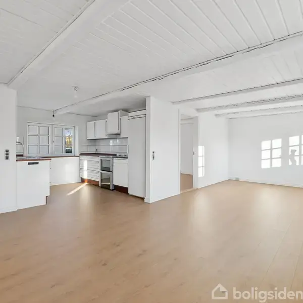 Rummeligt køkkenalrum med trægulve og hvidmalede bjælker. Køkkenet har hvide skabe og rustfri hvidevarer. En stor tom stue med hvide vægge og flere vinduer, der giver naturligt lys.