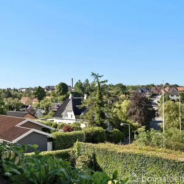 Huse med tage i forskellige farver ses spredt blandt frodige grønne træer og buske, under en klar blå himmel i et forstadsområde.