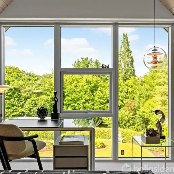 Et skrivebord med en lampe og dekorationer står i et rum med store vinduer, der åbner op til en udsigt over en frodig have med træer og buske.