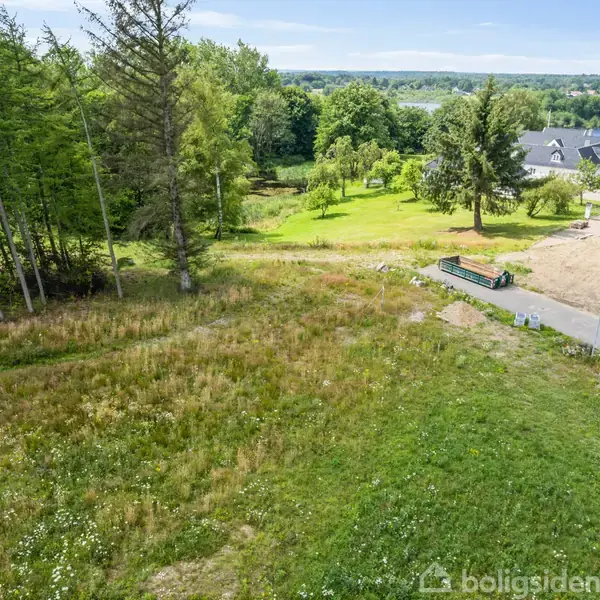 Græsplæne strækker sig over et åbent område ved siden af en vej med en hvid varevogn og moderne bygninger, omgivet af frodig skov og grønne træer, i nærheden af en sø.