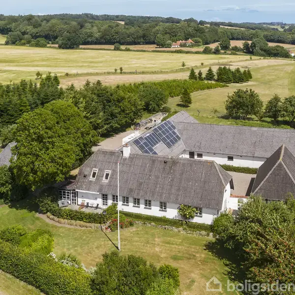 Et traditionelt stråtækt bondehus med solpaneler på taget ligger i en grøn, landlig omgivelser med træer og marker. Vejen løber langs huset, omgivet af buske og græs.