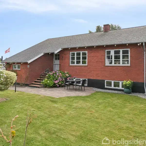 Rødt murstenshus med trappeopgang, omgivet af velplejet græsplæne og blomsterbede. Terrasse med havemøbler. Dannebrog er hejst i baggrunden under en blå himmel.