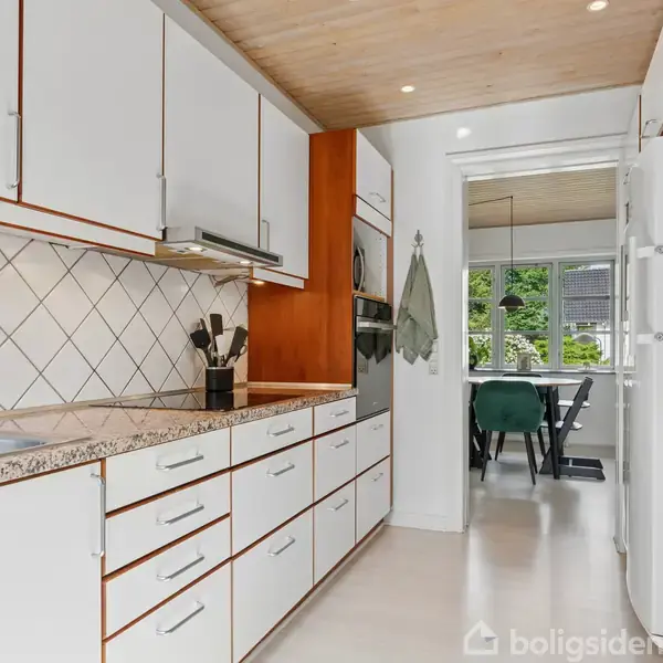 Et moderne køkken med hvide skabe og skuffer, marmorerede bordplader og knive på en magnetisk holder. En spiseplads er synlig gennem en døråbning, omgivet af grønne planter udenfor.