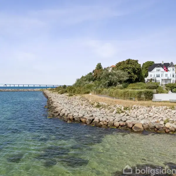 En hvid bygning står stille ved kysten på en klippefyldt strand, omgivet af grønne træer. Til venstre strækker en lang bro sig over det klare blå vand under en solrig himmel.