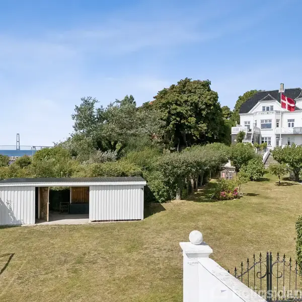 Hvid bygning med dannebrogsflag står ved havet. En stor have strækker sig ned mod en lille hvid bygning. Broen ses i baggrunden under en klar blå himmel.