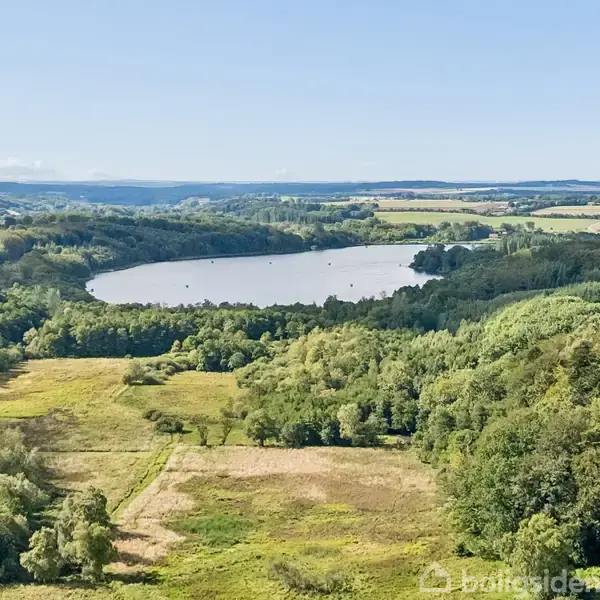 Sø omgivet af tæt skov og åbne marker, set fra et højt perspektiv. Landskabet er frodigt og grønt under en klar blå himmel.
